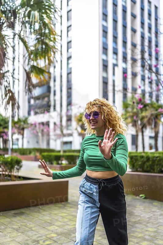 Energetic young woman with curly hair dancing in a modern city setting, surrounded by tall buildings and palm trees, expressing joy and freedom