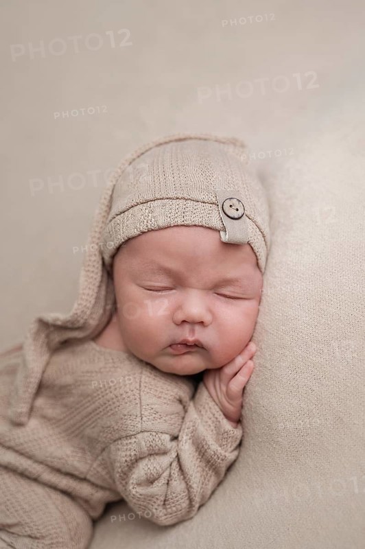 A serene newborn baby peacefully sleeps while dressed in a soft, beige knit outfit, complemented by a matching hat with a cute button. The scene exudes warmth and tranquility