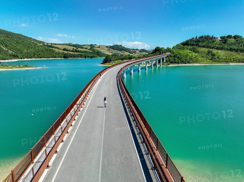 A young woman walks alone on a picturesque bridge surrounded by lush greenery and turquoise waters in the Italian countryside, highlighting nature's serene beauty