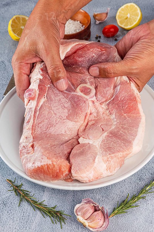 Raw pork shoulder being seasoned with garlic, lemon, salt, and herbs on a white plate. Hands holding the meat, ready for cooking, with spices displayed on a textured surface