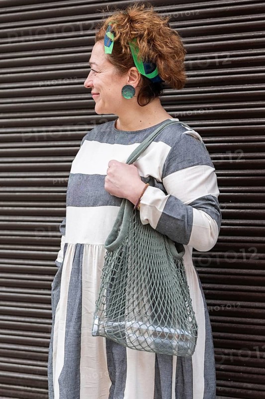 A woman in a stylish, grey and white striped dress stands confidently with her eco friendly mesh bag. She wears a colorful headband and statement earrings, showcasing sustainable fashion
