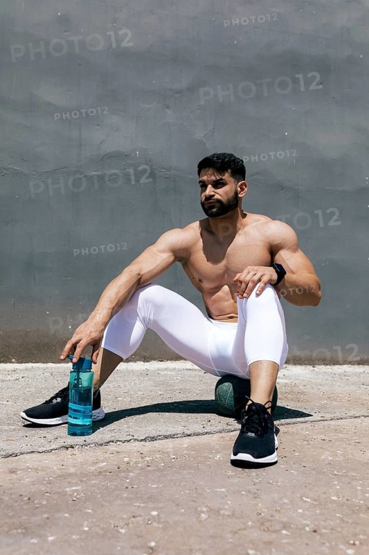 A fit man in white athletic leggings sits on a ball resting after an intense workout. He holds a water bottle, staying hydrated. Urban background enhances the sporty vibe