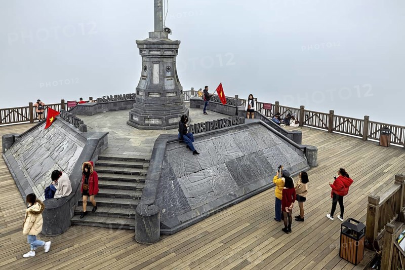 People on a platform in the mist, surrounded by railings and a flag. Stone structure with steps in the centre, The summit of Fansipan near Sa Pa in Vietnam