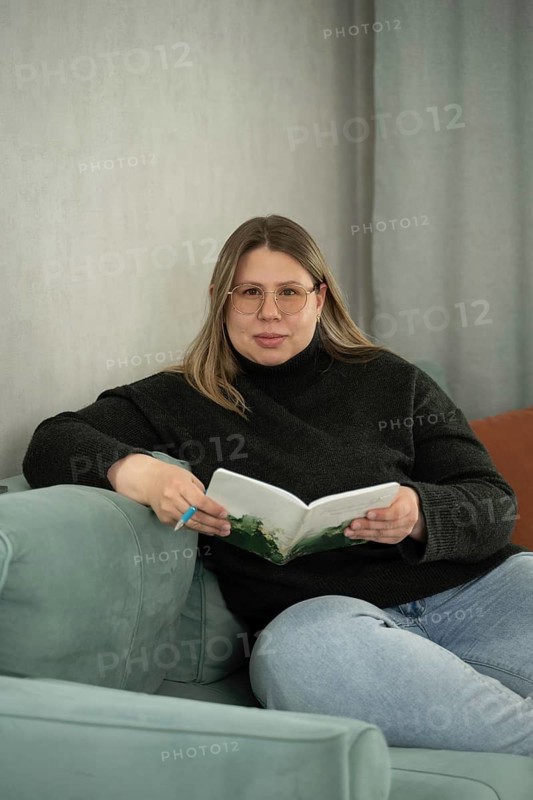 A woman in glasses relaxes on a soft sofa holding an open notebook and pen, dressed casually in a sweater and jeans. The calm setting and soft colors suggest a peaceful, reflective moment at home