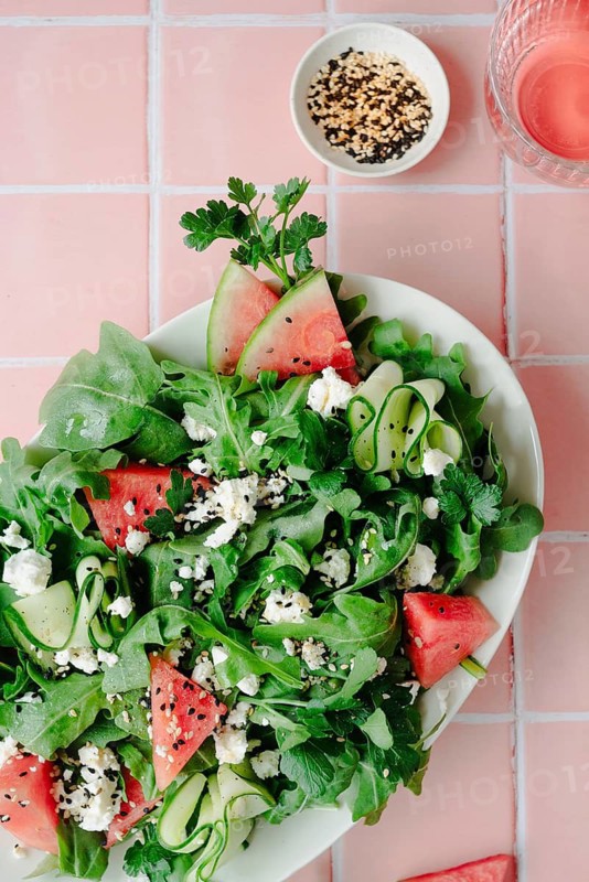 Top view of a nutritious salad combining ruccola, crisp cucumber slices, juicy watermelon pieces, and crumbled feta cheese, served on a white plate against a pink-tiled background