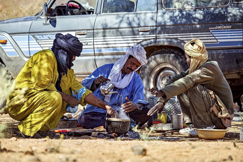 Three men in traditional dress prepare a meal outside, next to a jeep in a desert landscape, Tuareg sit together over tea in the Sahara in Algeria