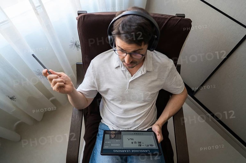 A man sits at home with headphones, intently studying Mandarin online using a tablet. The setup indicates a comfortable and focused learning environment