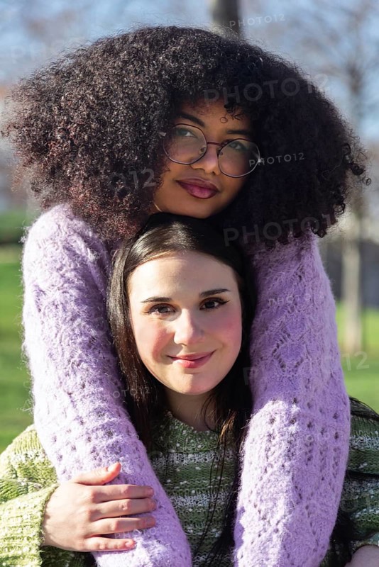 Two friends enjoy a sunny day outdoors, embracing warmly. One wears glasses and a purple sweater, and the other smiles beneath her friend's chin. A moment of closeness and joy