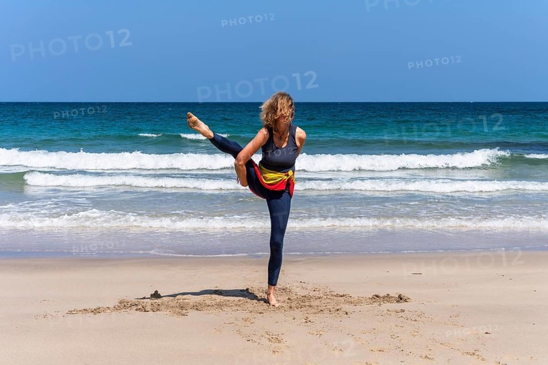 A woman doing yoga on a sandy beach by the sea. Svarga Dvijasana or bird of paradise. Treen Cove, Cornwall, England, Great Britain