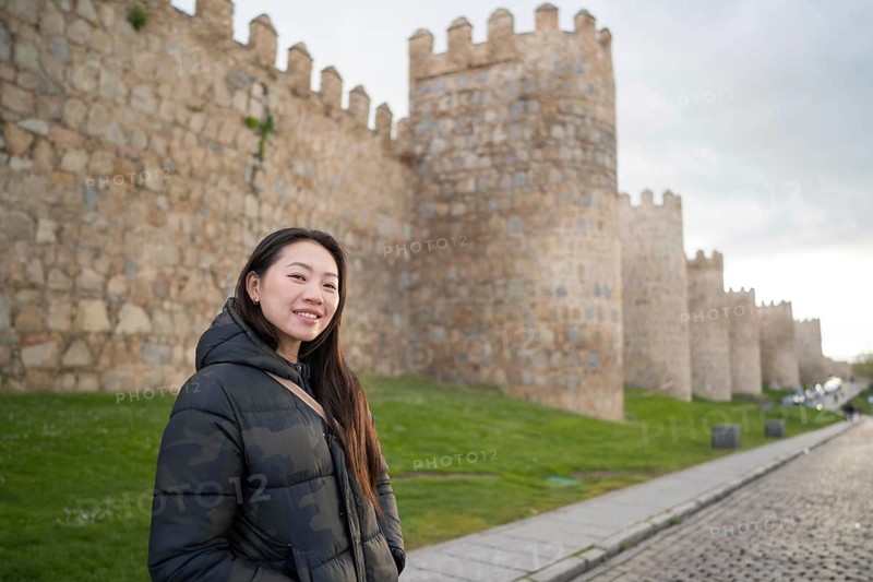 An Asian female tourist enjoys a scenic walk along the historic medieval walls of Avila, Spain. The ancient stone architecture contrasts beautifully with the green landscape