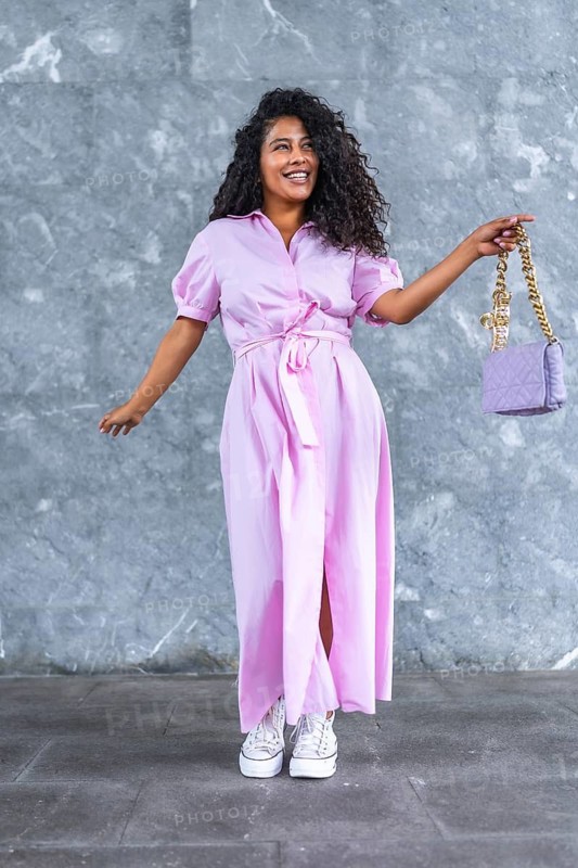 Vertical full length portrait of a happy elegant latin woman posing holding a chic purse against a grey urban wall