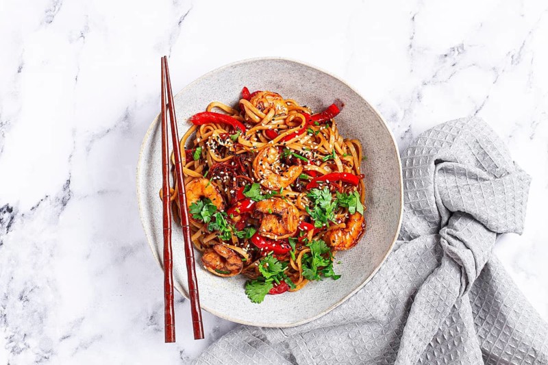 Udon noodles, shrimp with vegetables and fresh herbs, in a bowl with chopsticks, light background, homemade, no people