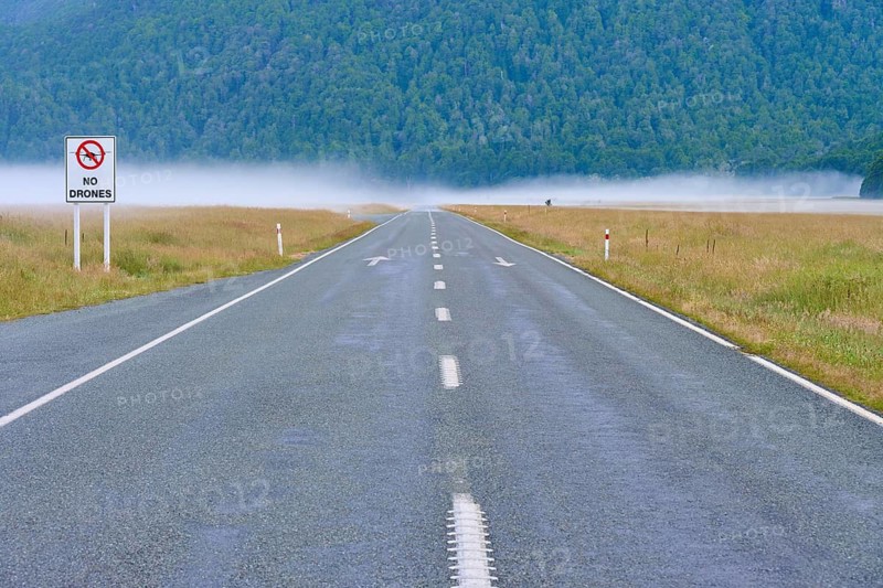 Straight tarmac road in foggy landscape with drone prohibition sign on the side, summer, Eglinton Valley Viewpoint, Milford Sound, Te Anau Downs, Fiordland National Park, South Island, New Zealand