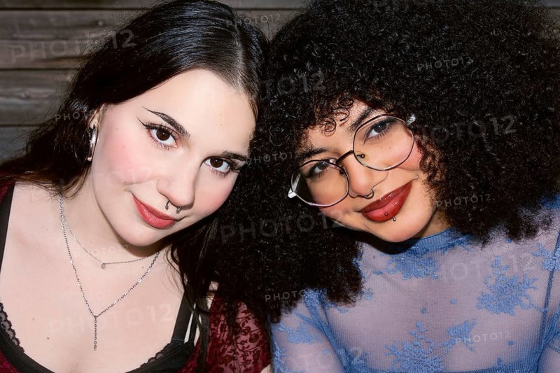 Two Gen Z friends, a Caucasian girl with dark straight hair and a Hispanic girl with curly hair, smiling warmly as they pose closely together in front of a wooden backdrop