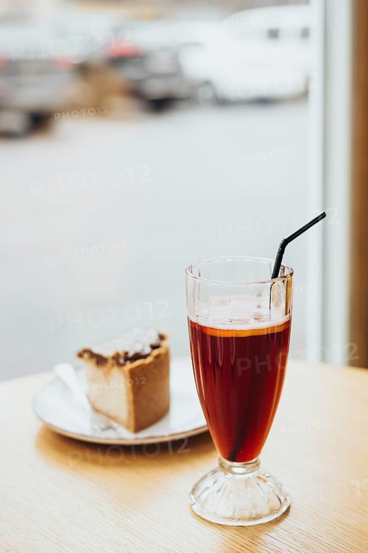 A glass of iced tea with a straw and a slice of cake on a wooden table near a window, offering a relaxed ambiance in a cozy cafe setting