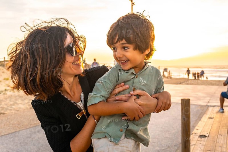 Mother and son are smiling and embracing on a wooden boardwalk at the beach during sunset, enjoying happy moments together during a summer vacation with golden light