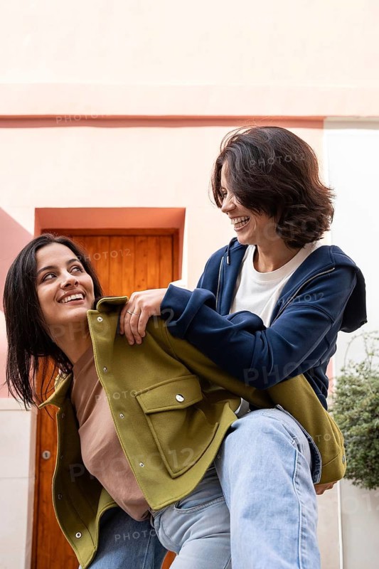 Two women enjoying a playful, joyful moment outdoors. They are dressed casually, smiling at each other, embodying happiness and connection against a pastel-colored backdrop