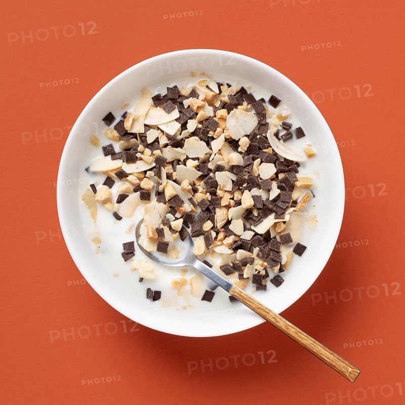 Top view of a white bowl filled with cereal, sliced almonds, and chunks of chocolate, all soaked in milk, set against a vibrant orange background. A wooden spoon rests in the bowl