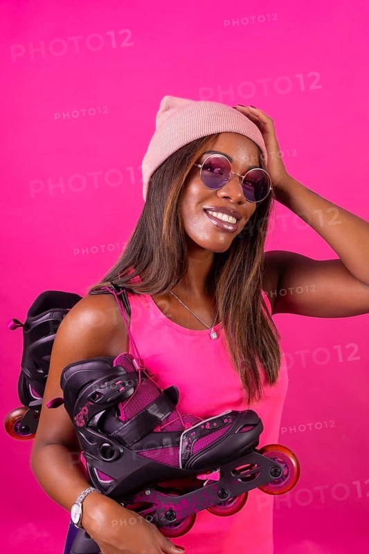 Vertical studio portrait with pink background of a cool african woman with urban hat and sunglasses carrying skates