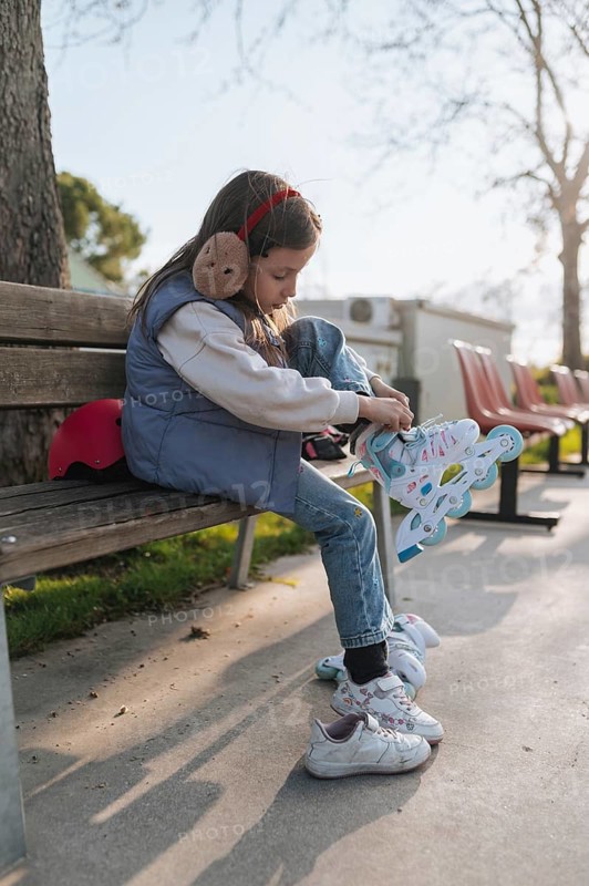 A young girl prepares to skate by fastening her roller skates while seated on a park bench with a red helmet beside her