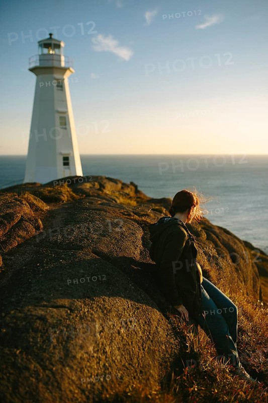 Side view of an unrecognizable woman sitting on rocks, overlooking the ocean near the Cape Spear Lighthouse in Newfoundland & Labrador, Canada during a serene sunset