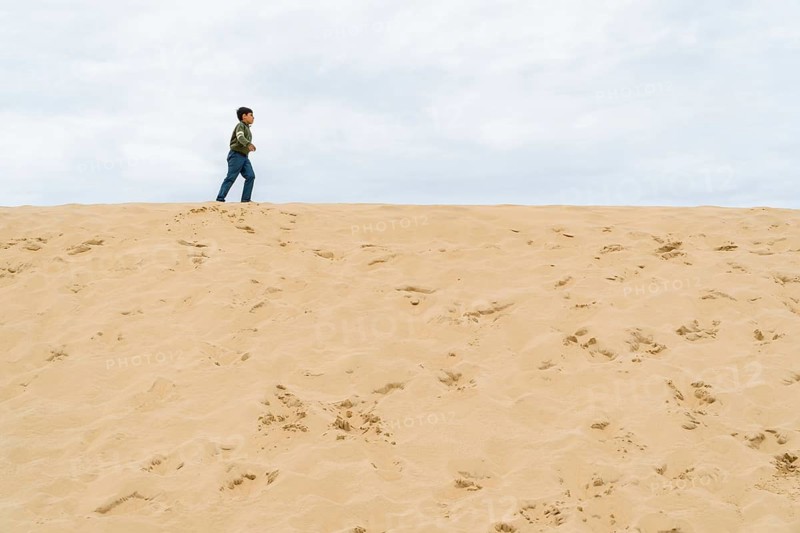 A young boy walks across the dune of Pilat under a cloudy sky in France. The minimal landscape emphasizes solitude and exploration, capturing a sense of adventure and tranquility