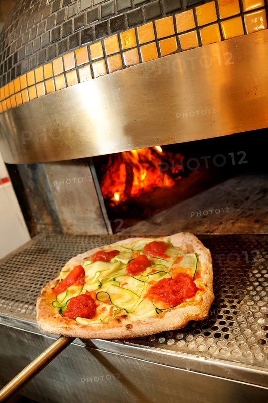 A freshly made pizza with toppings of tomato, pepperoni, and zucchini slices is baked inside a glowing wood-fired brick oven, highlighting the art of traditional pizza making