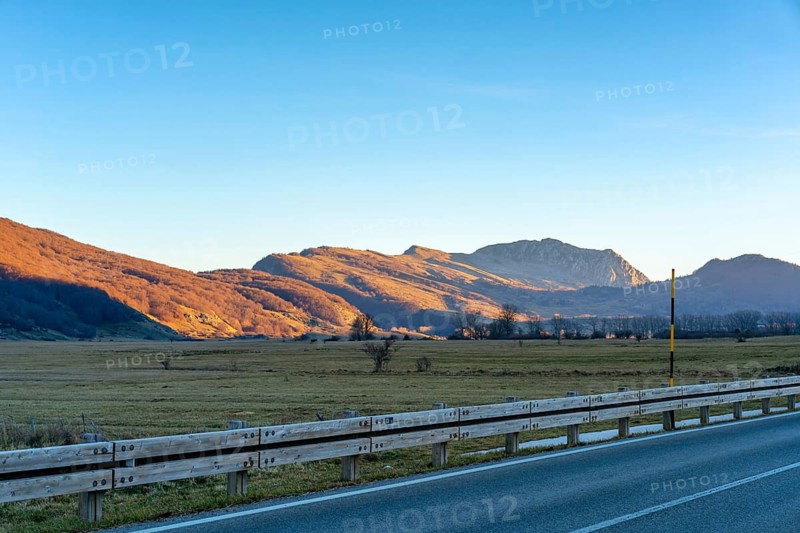 A serene mountain road in the Lazio region of Italy during sunset. The scene features golden-lit hills, open fields, and a quiet asphalt road with a wooden guardrail