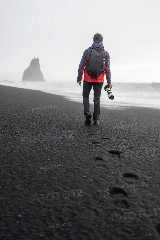 Back view of an unrecognizable photographer with a backpack and camera strolls along the misty shores of Reyinjajara Beach in Iceland, leaving a trail of footprints on the unique black sand