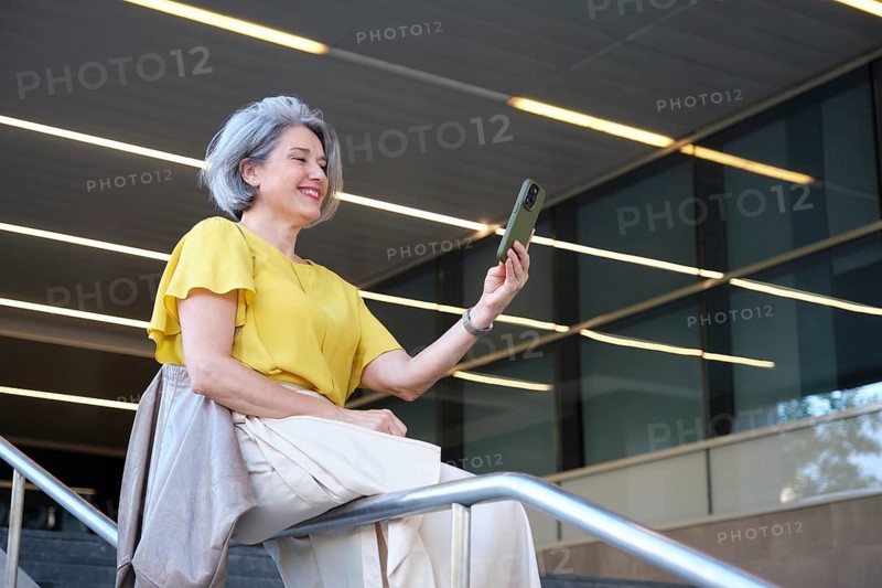 A cheerful mature woman in a yellow blouse takes a selfie with her smartphone while sitting on a railing outdoors. She is smiling and enjoying the moment in a modern setting