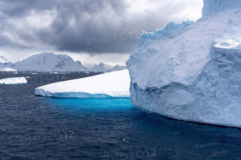 Table icebergs float in the dark Southern Ocean, turquoise underwater edge visible, snow-capped mountain range in the background under cloudy sky, Antarctic Peninsula, Antarctica