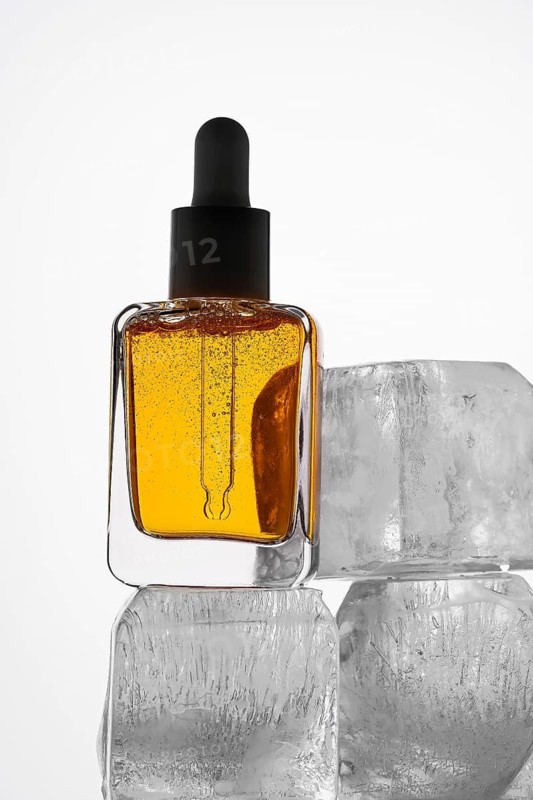 A vibrant orange Vitamin C serum in an elegant glass pipette bottle is placed on clear ice cubes, highlighting freshness and beauty in skincare products