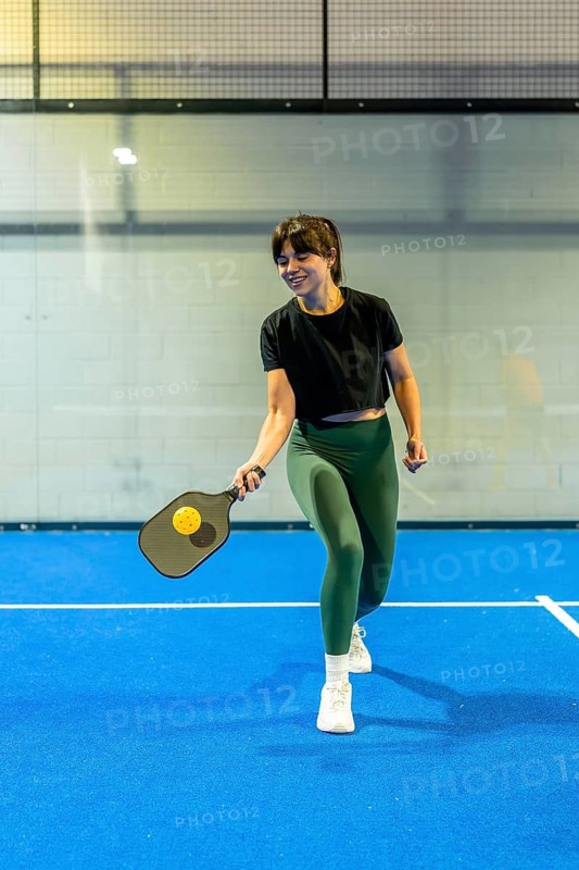 Young athletic woman actively participating in a pickleball game on a vibrant blue indoor court, holding a paddle and preparing to hit the yellow ball