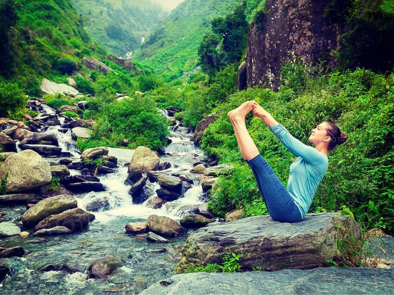 Yoga exercise outdoors, panorama of woman doing Ashtanga Vinyasa Yoga balance asana Ubhaya padangusthasana Big Double Toe Yoga Pose at tropical waterfall in Himalayas in India. Panoramic image. Vintage retro effect filtered hipster style image