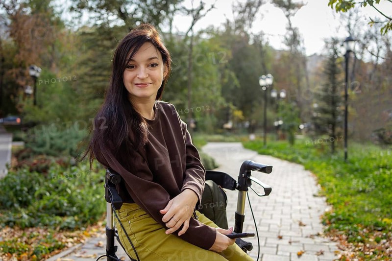 A woman with cerebral palsy with long dark hair sits comfortably on her walker in a sunlit park, holding a phone. She smiles at the camera, surrounded by lush greenery