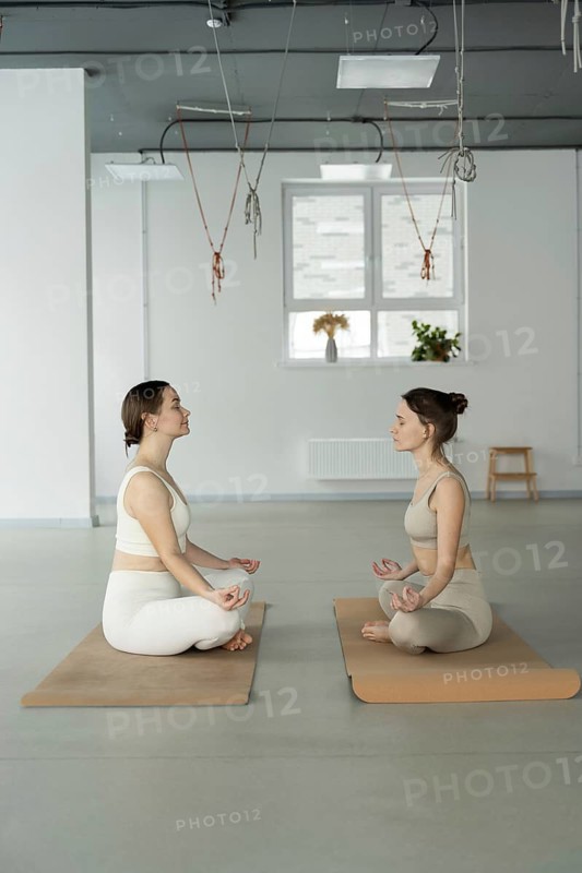 Two women sit cross legged on yoga mats, practicing mindfulness meditation in a serene studio setting. Natural light streams through the window, enhancing tranquility