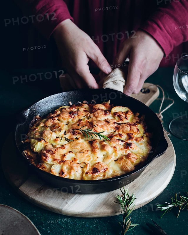 Hands arrange a sizzling dish of potato gratin topped with herbs in a black skillet. The wooden board enhances the rustic culinary aesthetic of this delicious meal
