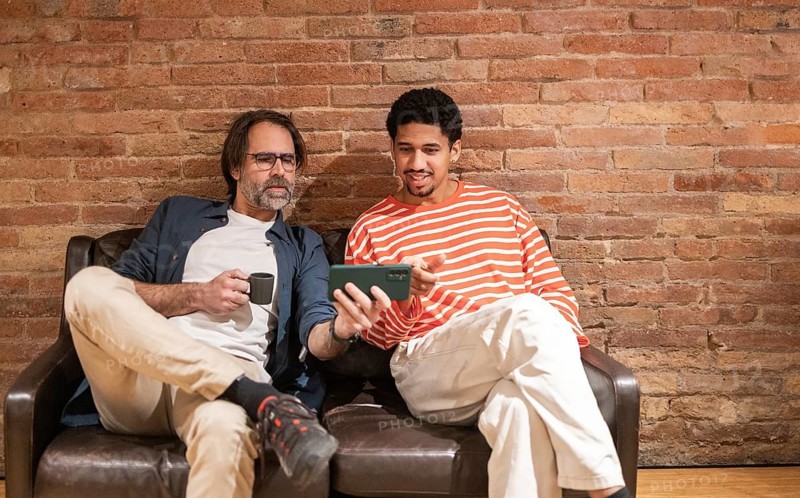 Two men relax on a leather sofa in a cozy coworking space, sharing a joyful moment while browsing content on a smartphone with a rustic brick wall as a backdrop