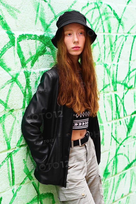 A young woman with long red hair and a black bucket hat poses against a vibrant green graffiti wall She wears casual street wear, exuding a modern and trendy vibe