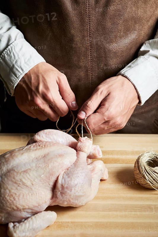 A butcher skillfully ties a raw chicken with twine on a wooden table, demonstrating traditional techniques. Hands are focused on ensuring precision in preparation