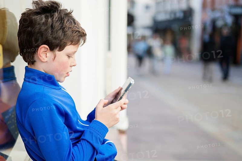 A boy in a blue jacket is deeply focused on his smartphone while leaning against a wall in a bustling city environment. The background showcases people walking past