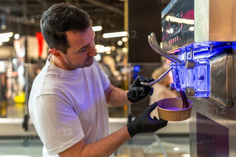 Man wearing black gloves serving purple soft serve ice cream into a disposable paper cup from a modern self service machine, preparing a dessert in a bright commercial setting