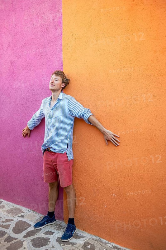Young man in front of pink and orange house wall with backpack, colourful houses on the island of Burano, Venice, Veneto, Italy