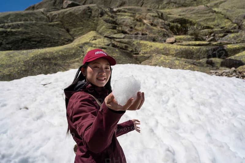 An Asian woman enjoys her winter adventure in the Sierra de Gredos, Spain. She's dressed warmly, holding a snowball, with rocky, snow-covered peaks in the backgroun
