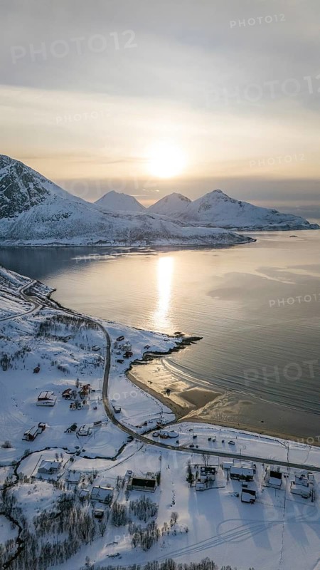 A stunning aerial view captures the serene beauty of a Nordic landscape, showcasing snow-covered mountains and a tranquil coastline kissed by the soft glow of the sun, Kvaløya, Troms, Norway