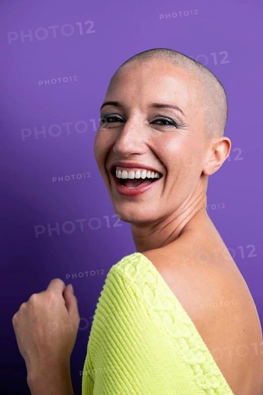 A vibrant portrait of a middle aged woman with a shaved head, smiling brightly against a purple background. The image captures the essence of joy, confidence, and style