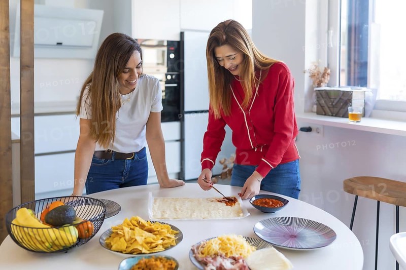Two cheerful women spreading tomato sauce on pizza dough and enjoying cooking together with various ingredients in a modern kitchen