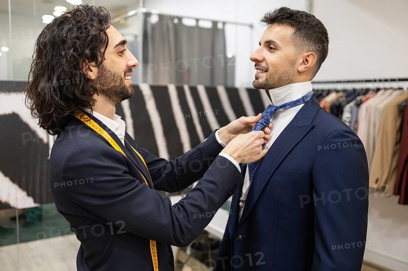 A tailor with a measuring tape around his neck carefully adjusts a customer tie in a suit store. The scene highlights professional service and attention to detail in men fashion