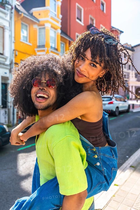 Young man giving piggyback ride to his girlfriend, both wearing sunglasses and smiling, enjoying a day out in the city