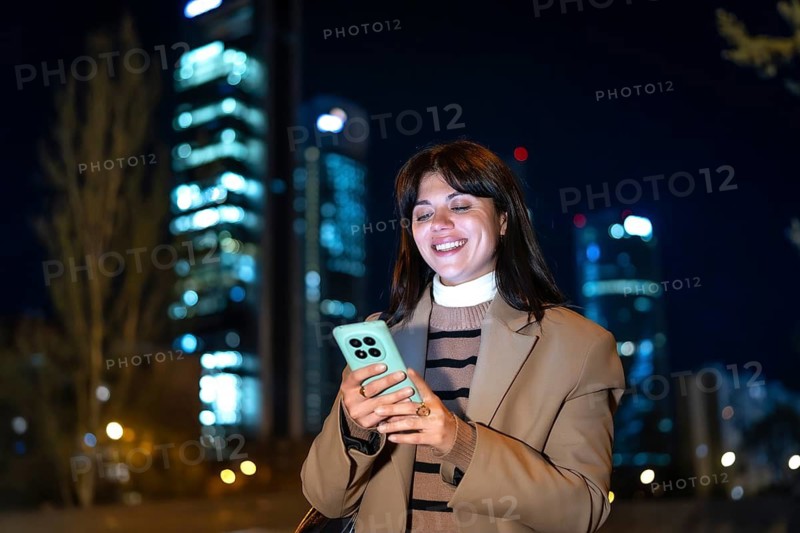Woman standing outdoors at night, smiling and concentrating on her smartphone, connecting with people and technology in an urban environment with illuminated buildings in the background